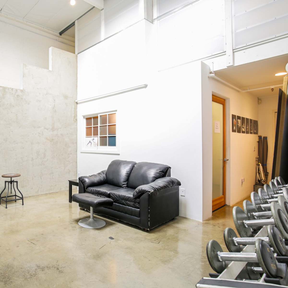 A black leather sofa and a small table are positioned near a concrete wall with stairs, adjacent to a row of gym weights in a well-lit space.