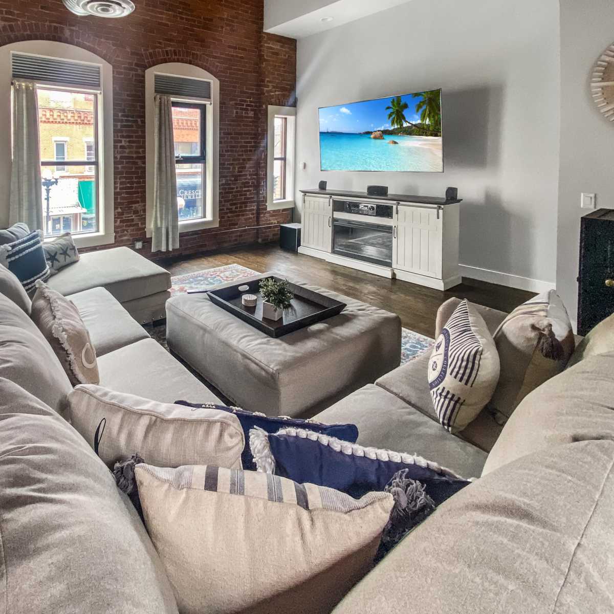 The living room features a large, U-shaped sofa, a coffee table in the center, and a television mounted on the wall, with exposed brick walls and large windows showing a view of the street outside.