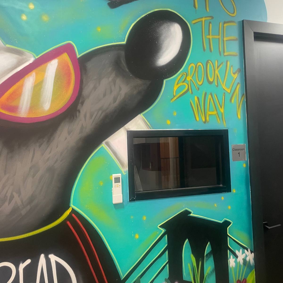 A colorful mural featuring a cartoon character wearing sunglasses and a cap is painted on a wall beside a window labeled "Classroom 1."