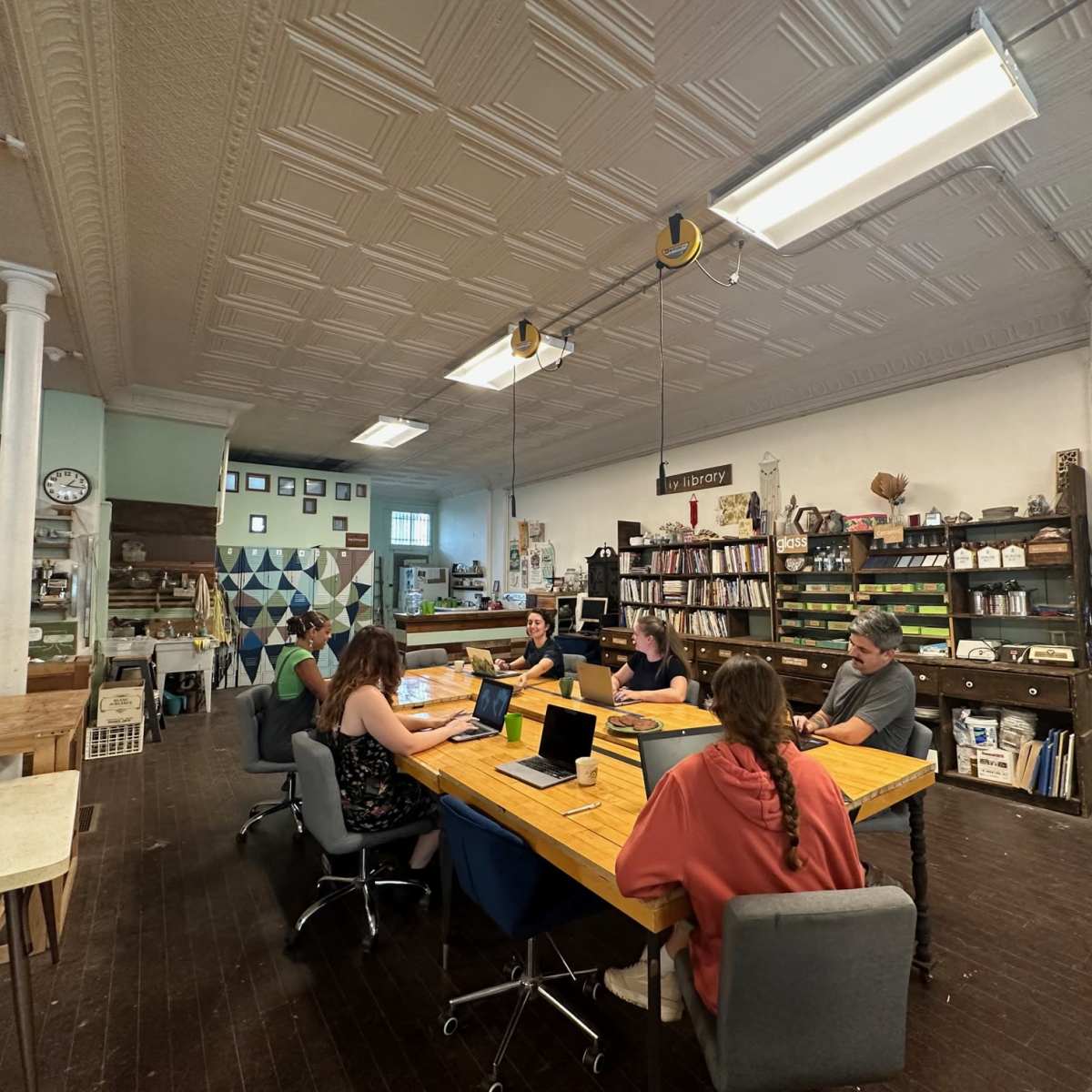 A group of six people are seated at a large wooden table in a workshop space, working on laptops while surrounded by shelves filled with various craft materials.