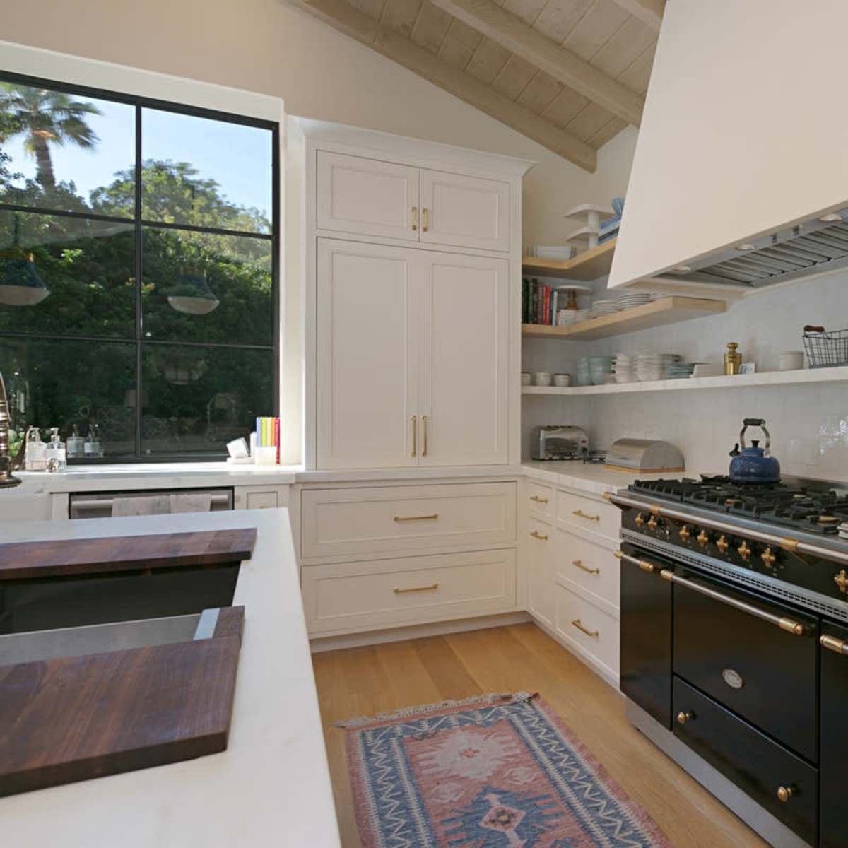 A modern kitchen featuring white cabinetry, a large black stove, open shelving with dishware, and a window providing natural light.