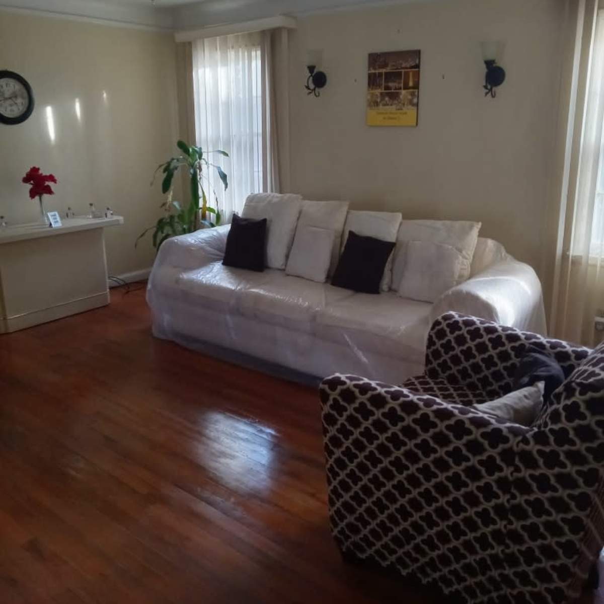 The living room features a white sofa with black cushions, a patterned armchair, a small table, and a houseplant against a backdrop of wooden flooring and cream-colored walls.