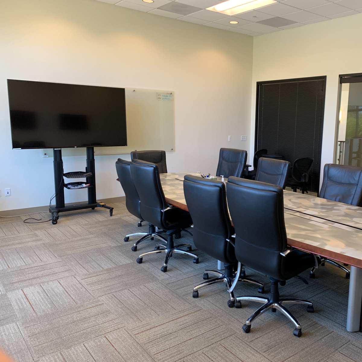 A conference room features a long table surrounded by leather chairs, a large television on a stand, and glass doors leading to another area.