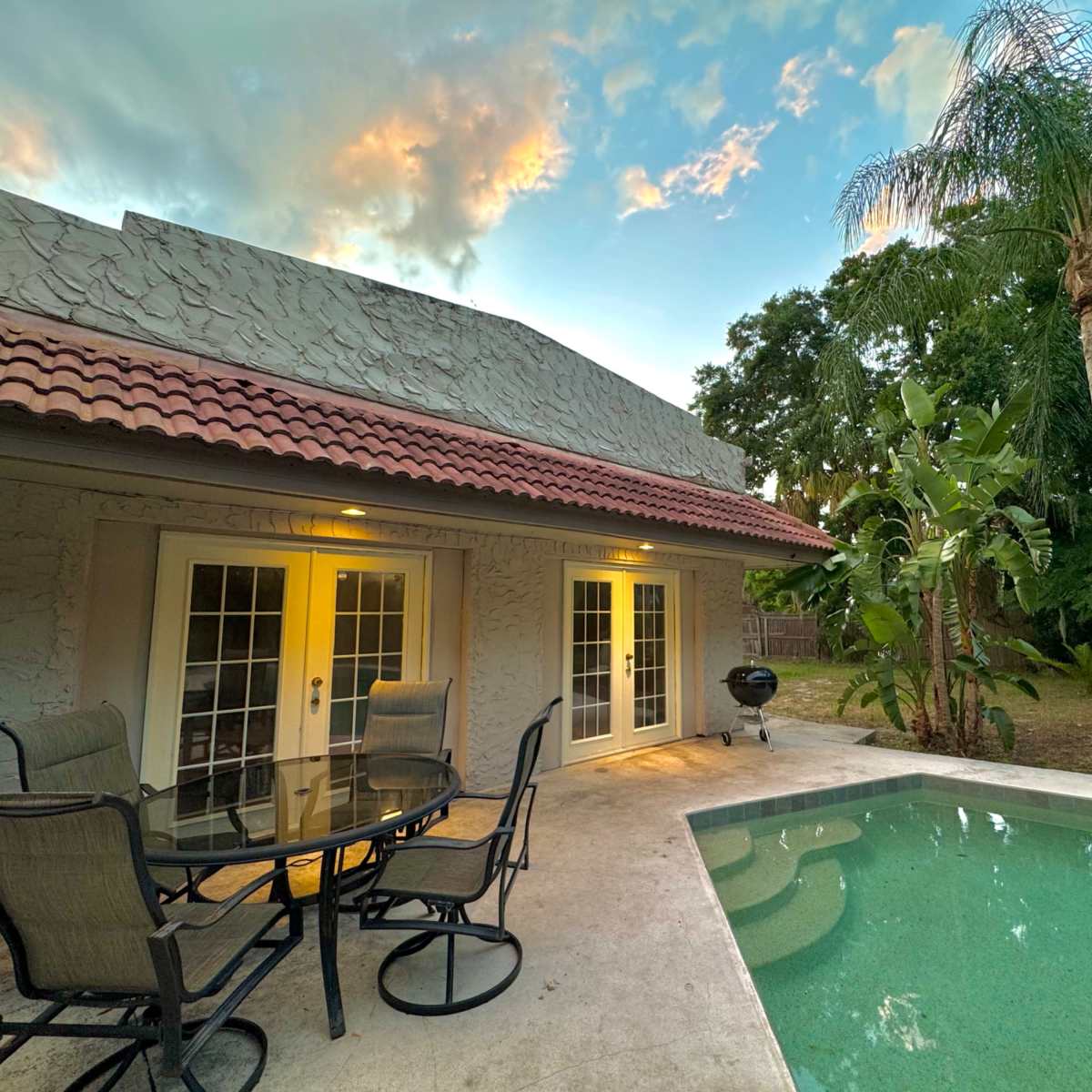 A patio area with a table and chairs overlooks a swimming pool surrounded by palm trees and tropical plants near a house at dusk.
