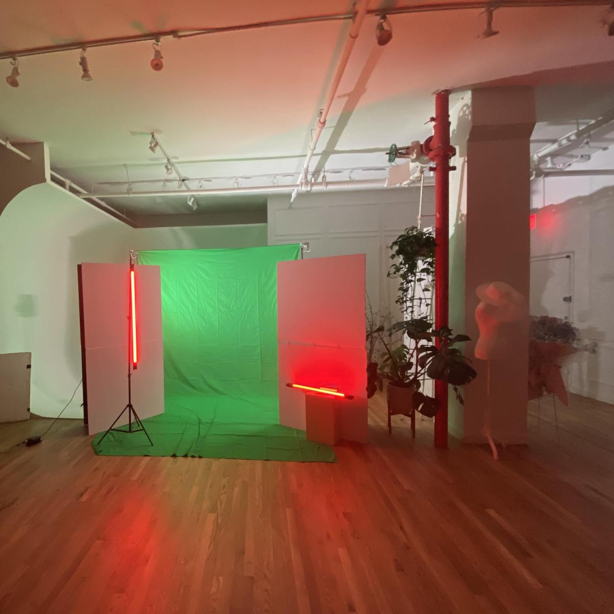 A studio space with a green backdrop, illuminated by red neon lights, surrounded by white partitions and plants.
