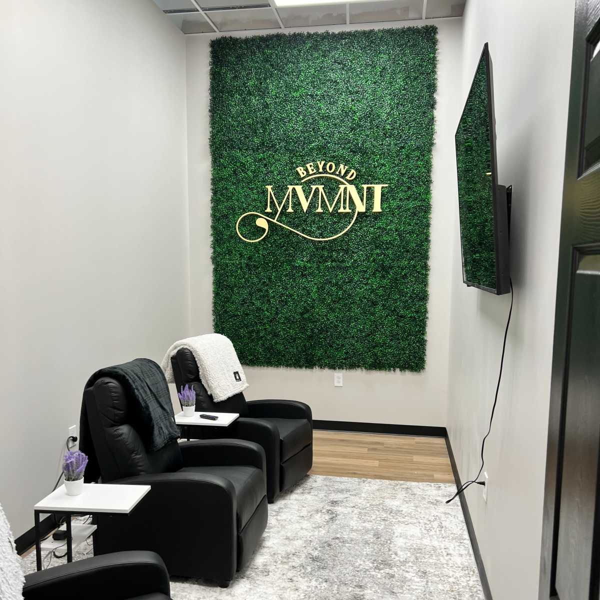 A small room designed for relaxation, featuring two black reclining chairs, a greenery wall with the words "BEYOND MVMT," and a television mounted on the wall.