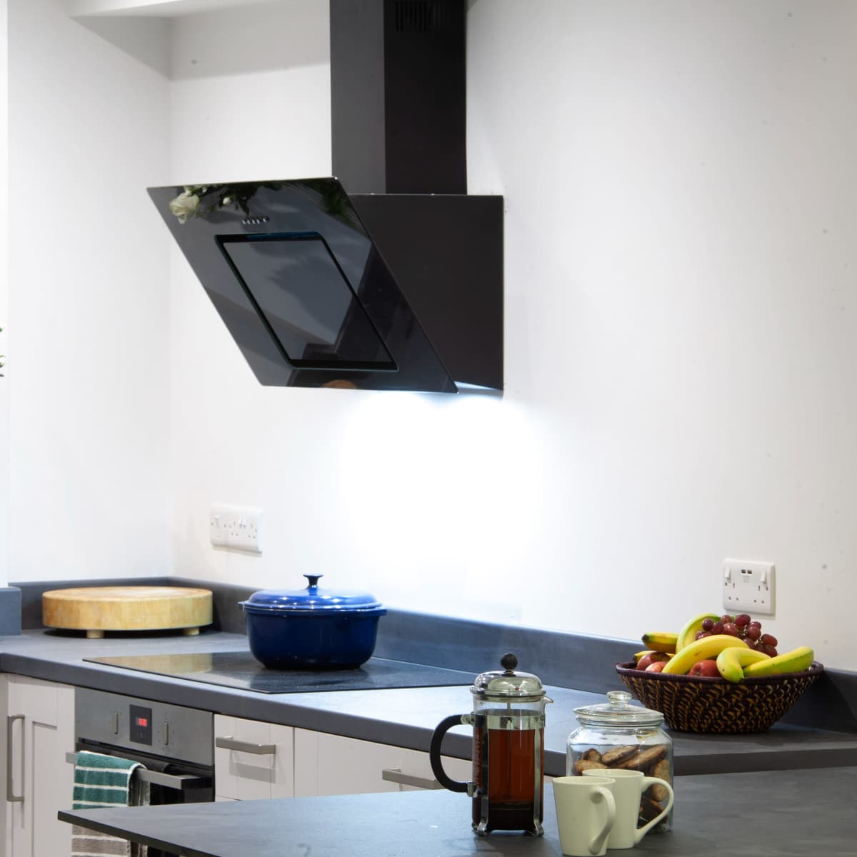 A modern kitchen features a black exhaust hood above a stove, with a blue pot on the countertop and a basket of fruit nearby.