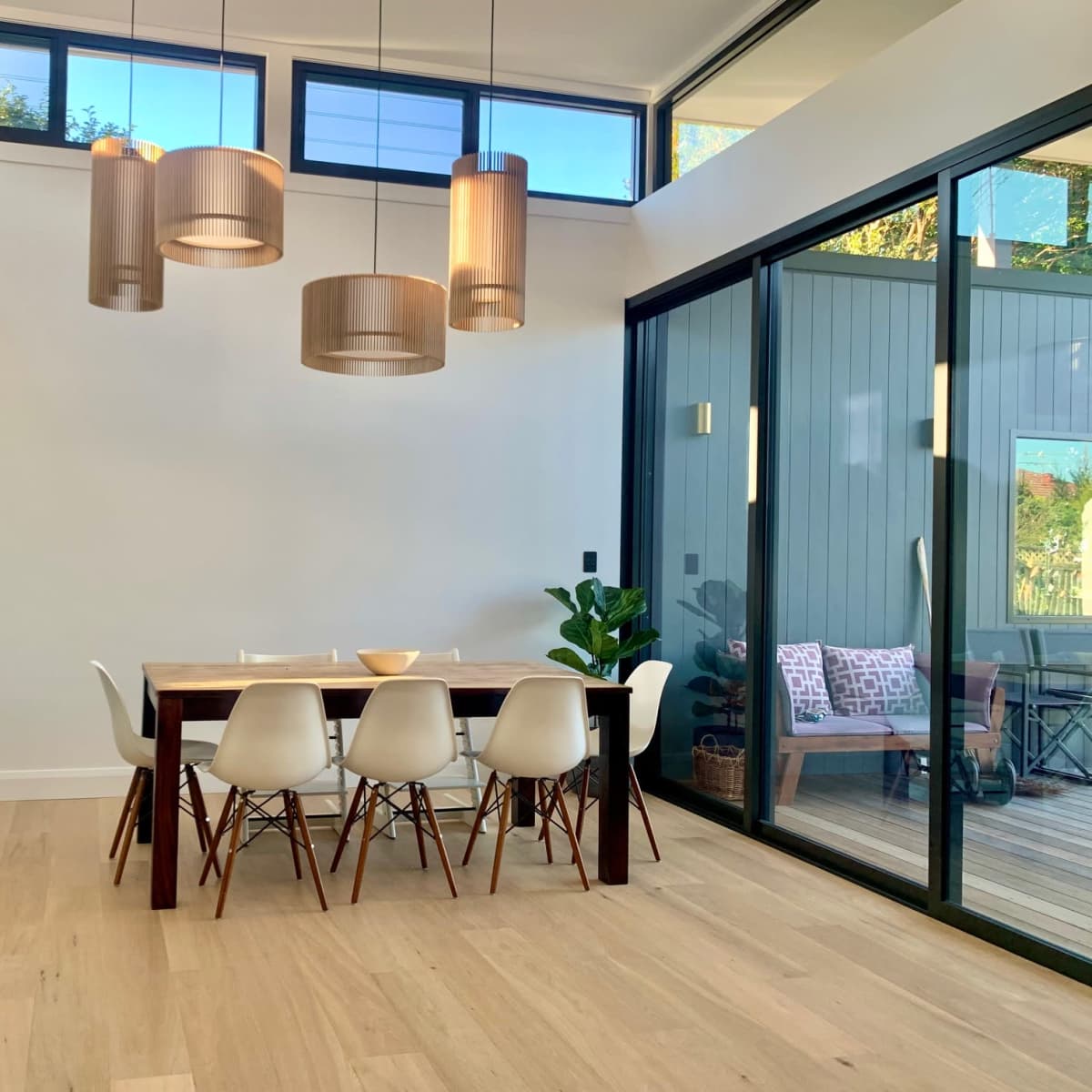 A modern dining area features a wooden table surrounded by white chairs, with pendant lights overhead and large glass doors opening to an outdoor space.