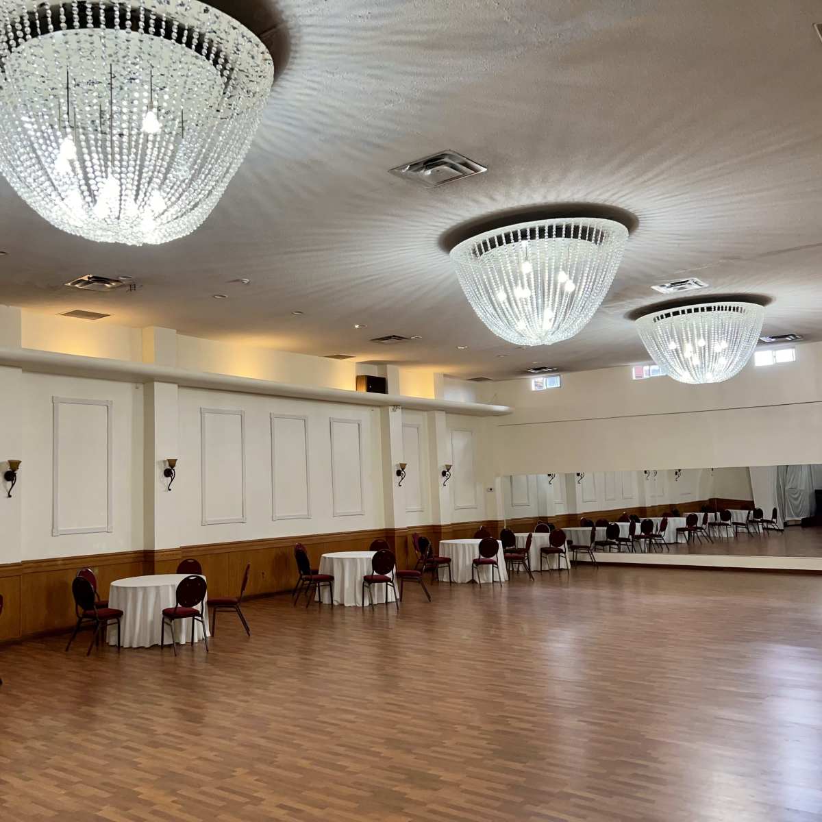 A large, empty banquet hall with two chandeliers, round tables draped in white cloth, and a mirrored wall.