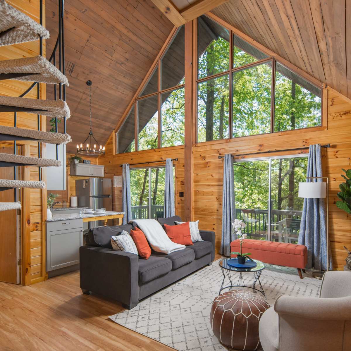 A wooden interior of a cabin with high ceilings, a spiral staircase, a living area featuring a gray sofa, a coffee table, and large windows overlooking greenery.