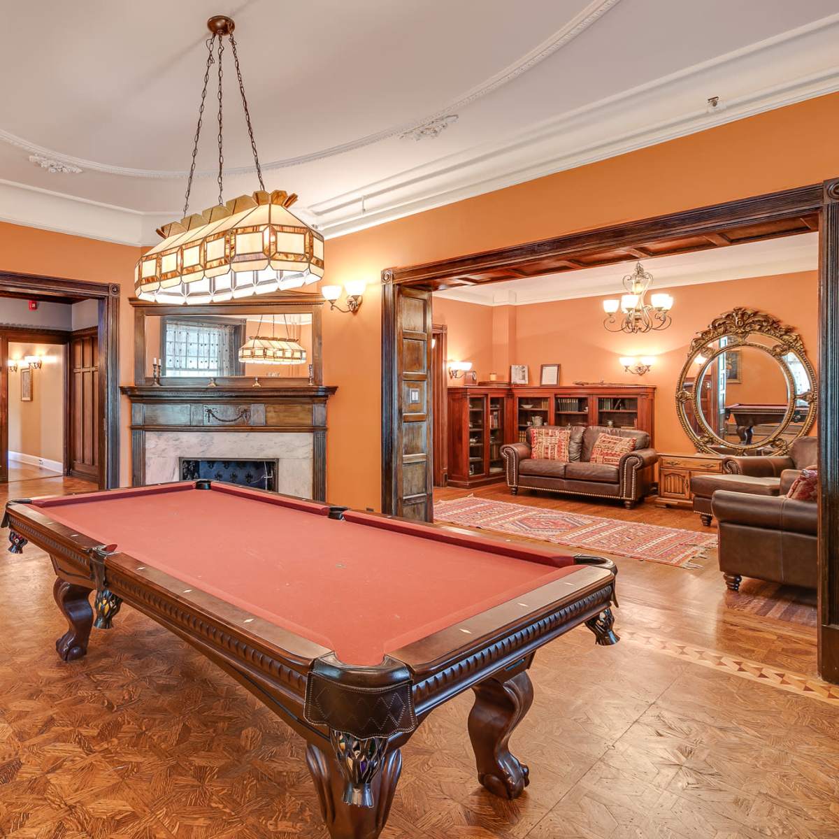 A room with a red billiards table centered under a chandelier, surrounded by walls painted orange and wooden accents.