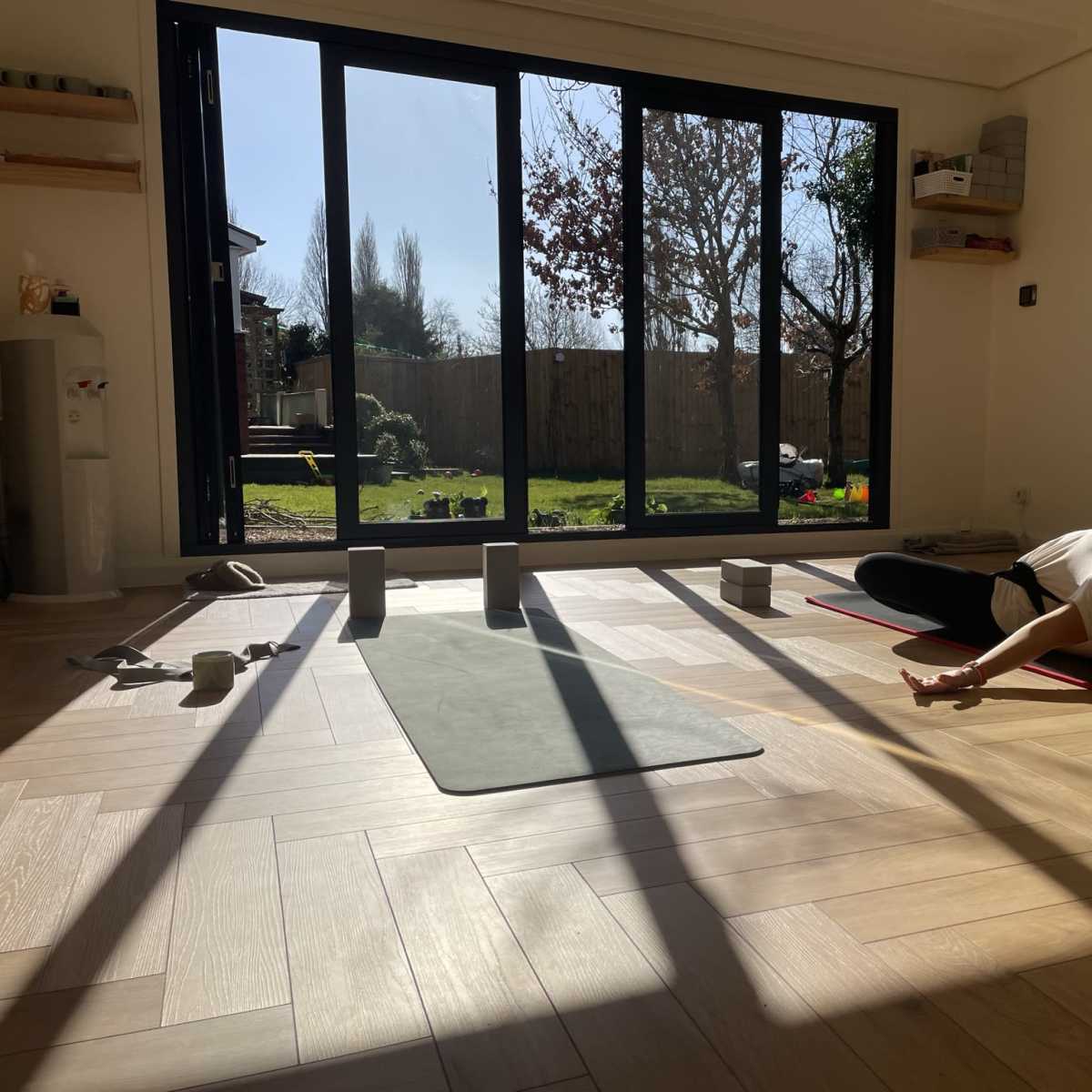 A person practices yoga on a mat in a sunlit room with large glass doors opening to a garden.