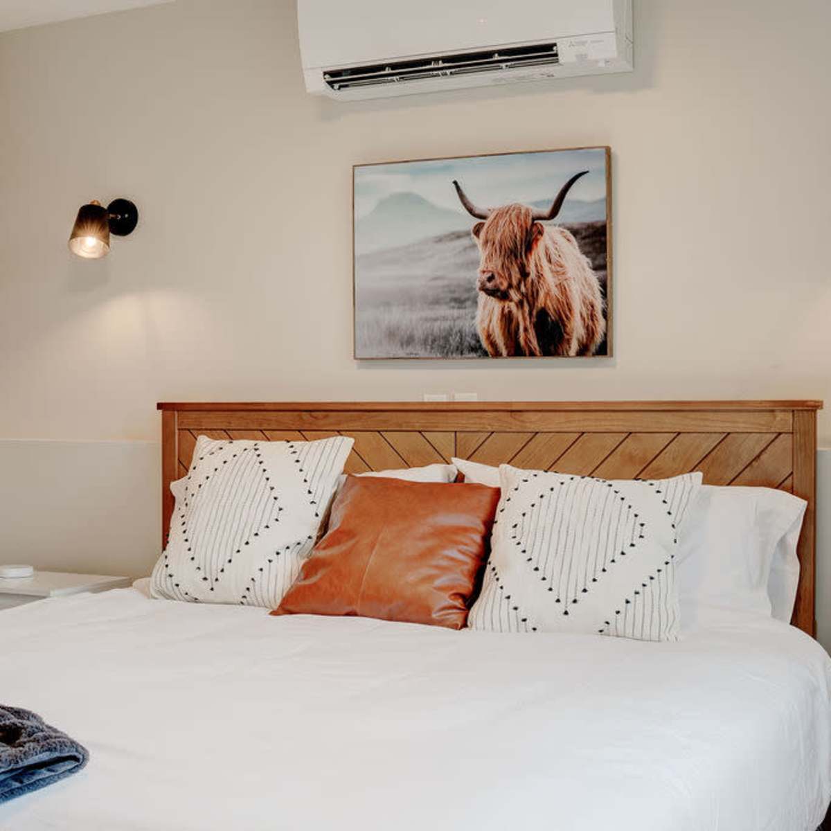 A modern bedroom features a wooden bed with decorative pillows, a wall-mounted air conditioner, and a framed animal portrait above the headboard.