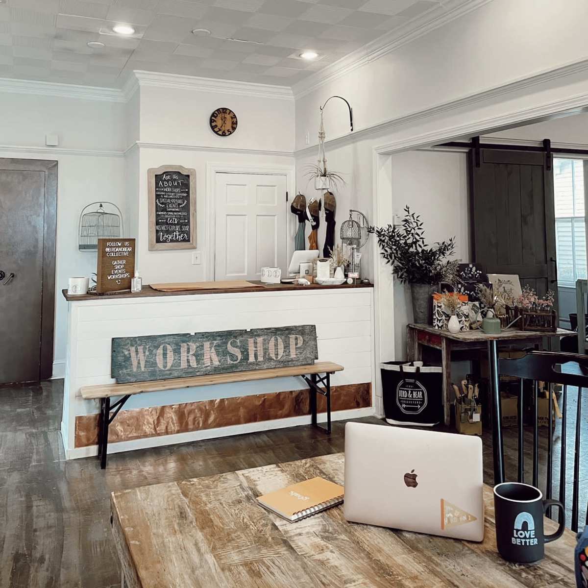 A cozy workshop reception area featuring a wooden counter, a sign that reads "WORKSHOP," and various decorative items like mugs, plants, and a laptop on a table.