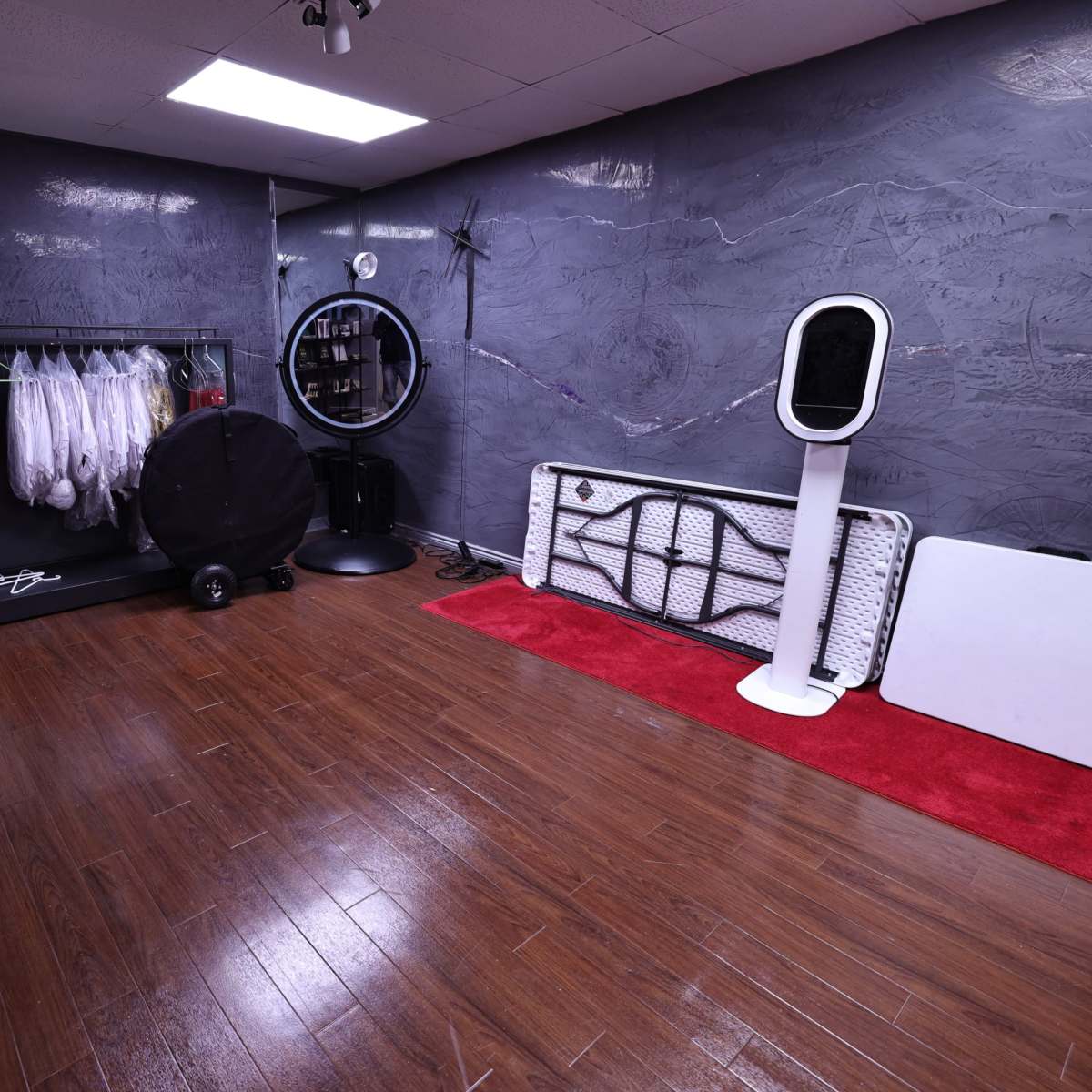 A room with wooden flooring, featuring a clothing rack with white garments, a circular mirror, and various equipment along the walls.