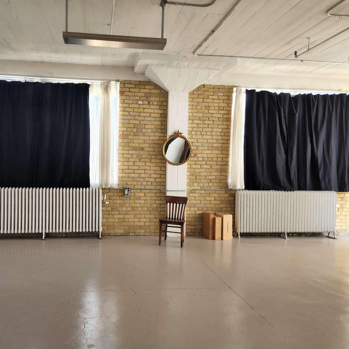 An empty room with exposed brick walls, a single chair, a mirror on the wall, and large windows covered by dark curtains.