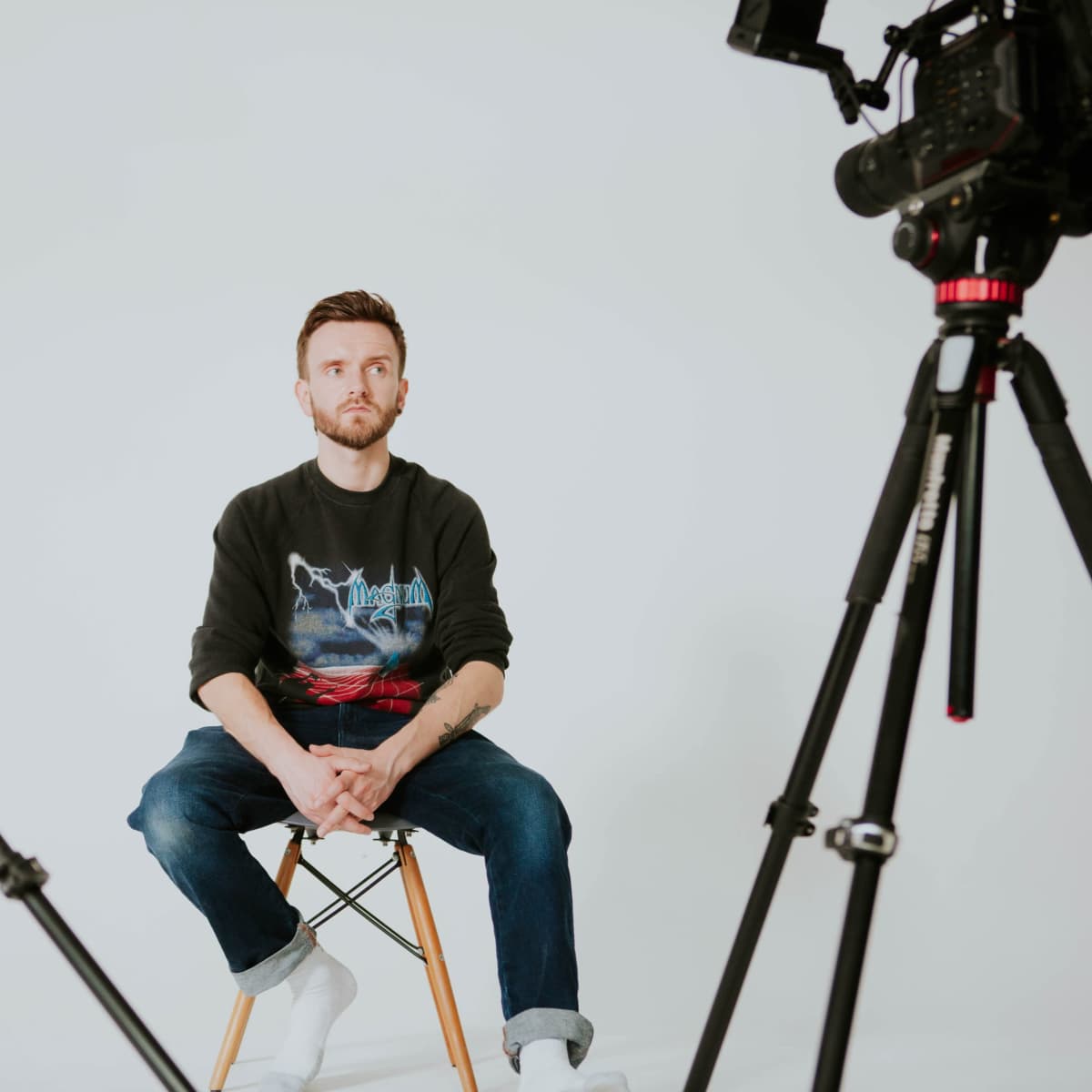 A man sits on a stool between two cameras on tripods against a plain white background.