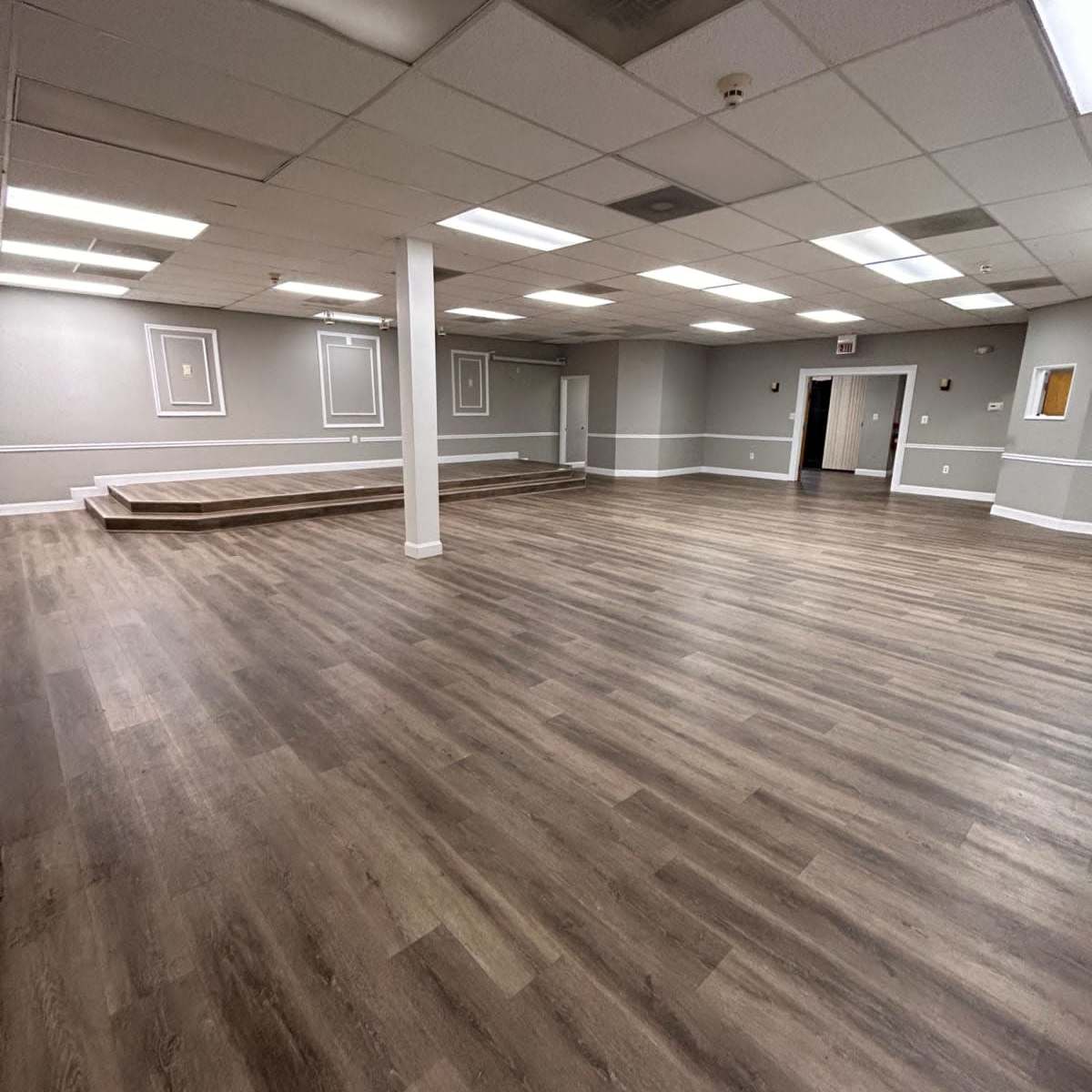 A spacious, empty room with wooden flooring, white walls, and a raised platform at one end.