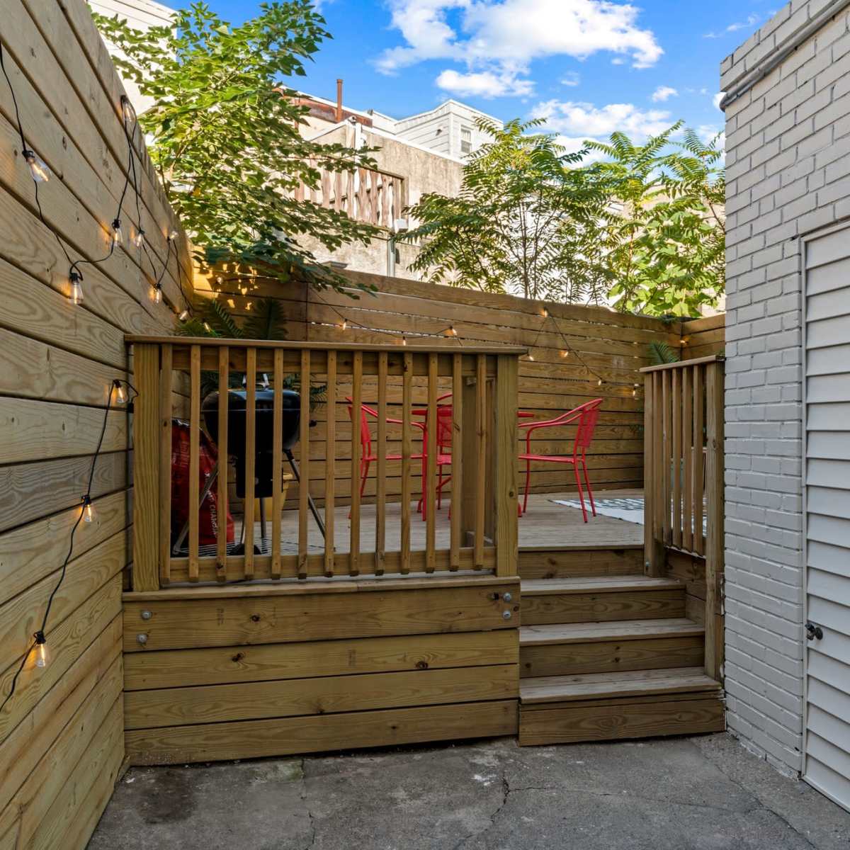 A small outdoor patio area with wooden decking, red chairs, and surrounding greenery, enclosed by wooden fences and a white brick wall.