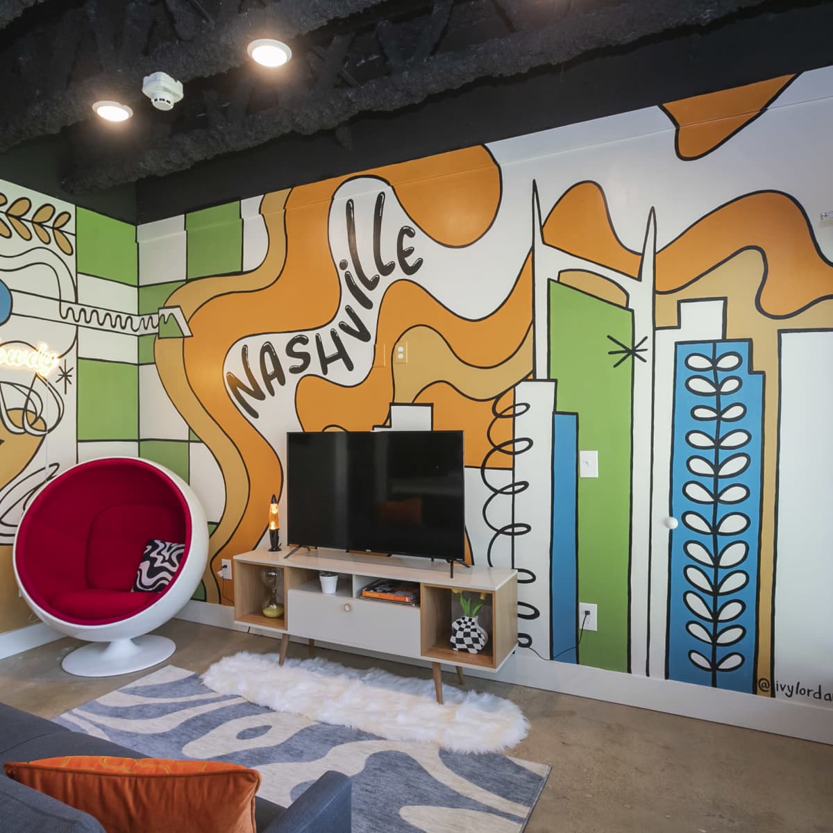 A modern living area featuring a brightly colored mural on one wall, a red egg-shaped chair, a television on a wooden stand, and a concrete floor.