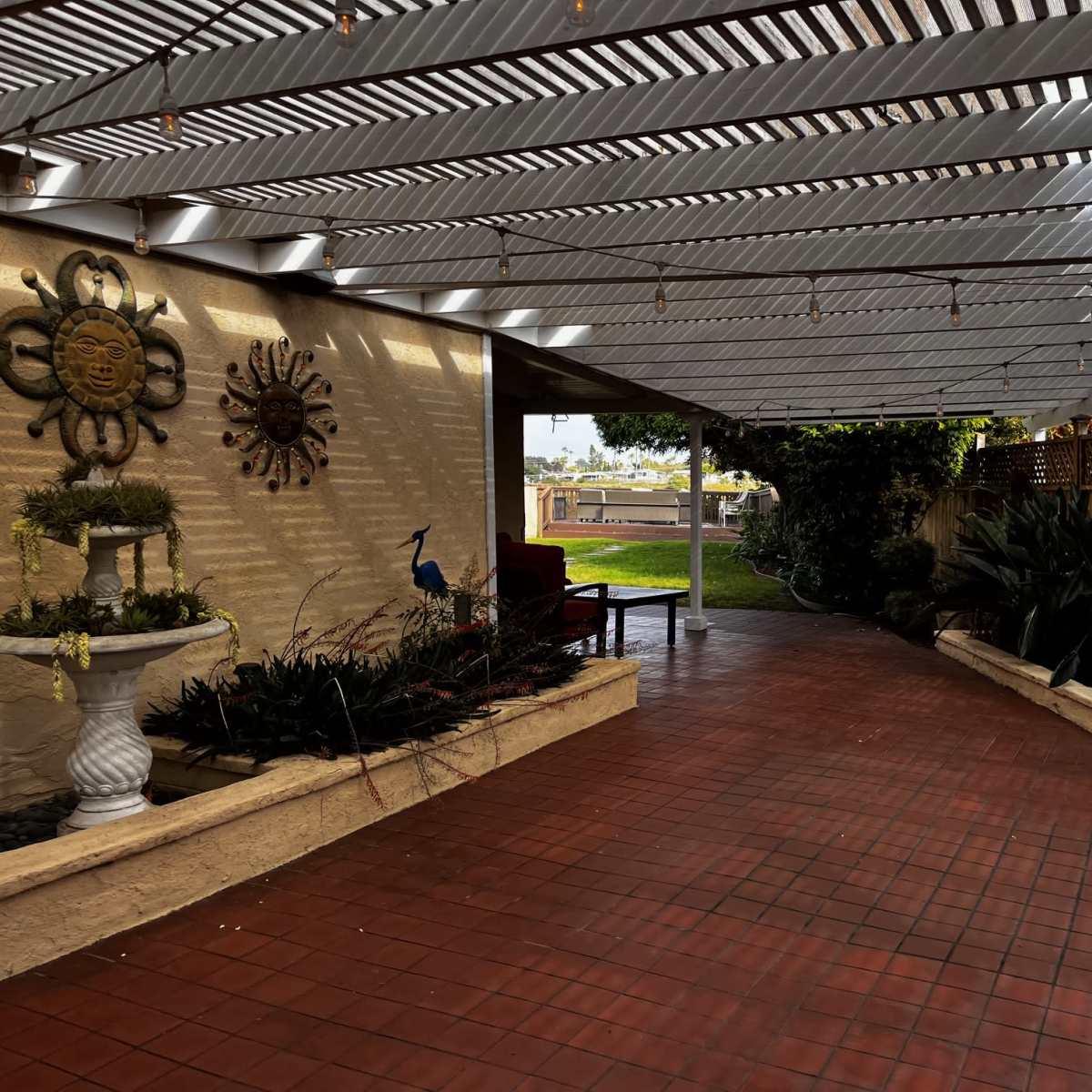 A covered walkway bordered by plants and decorative elements, leading to a grassy area beyond.