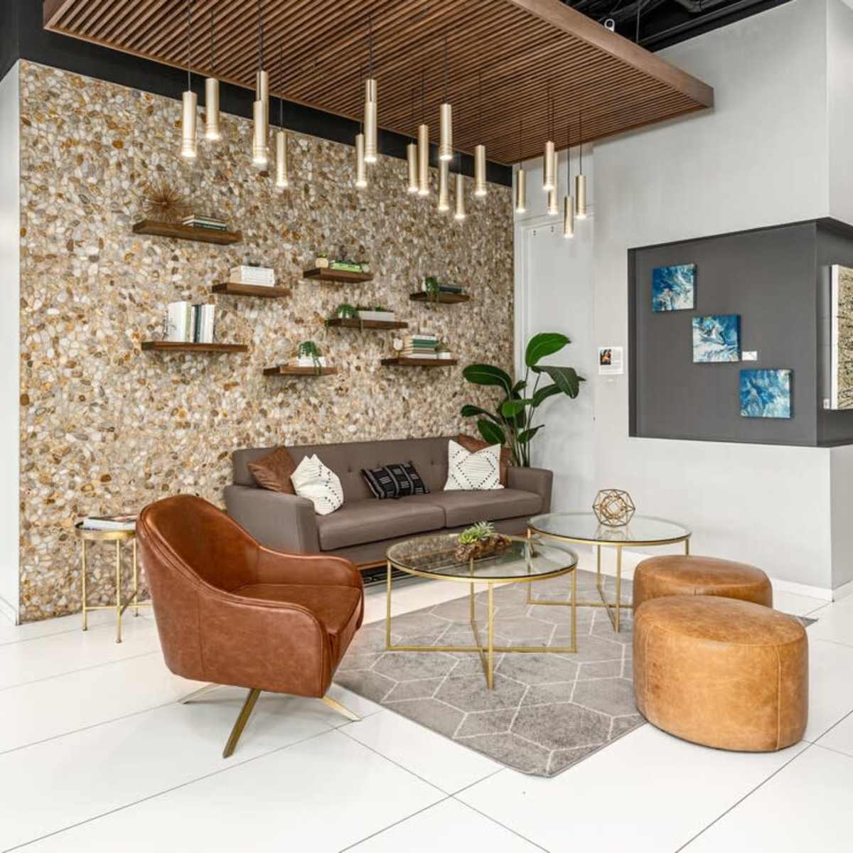 A modern lounge area with a brown sofa, decorative shelving on a textured wall, and gold accents, surrounded by circular wooden tables and greenery.
