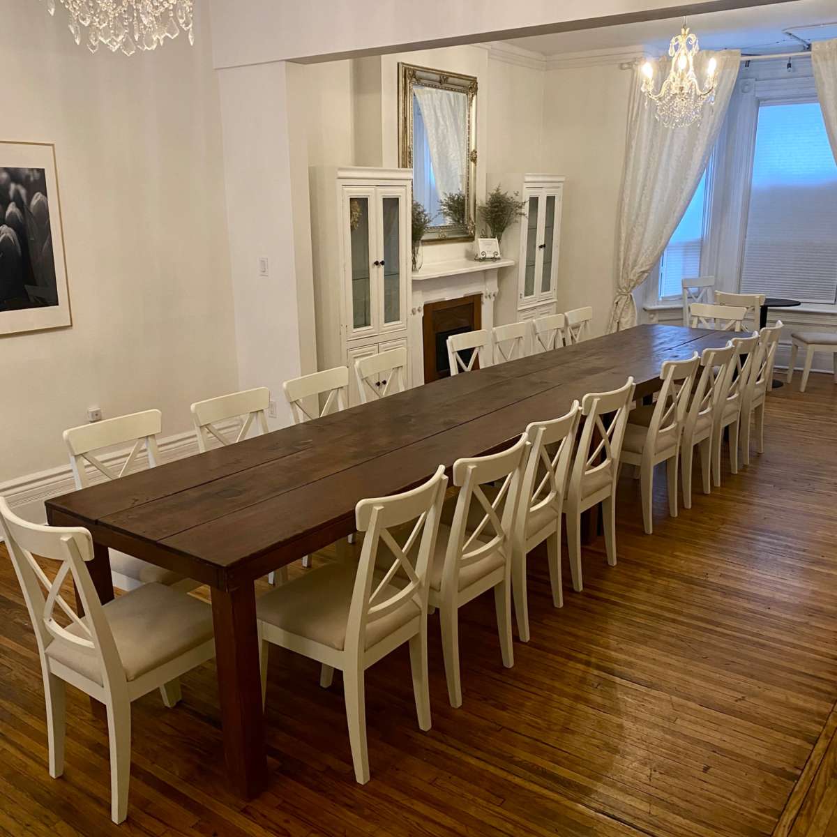 A long wooden dining table with white chairs is set in a well-lit room featuring chandeliers and large windows.
