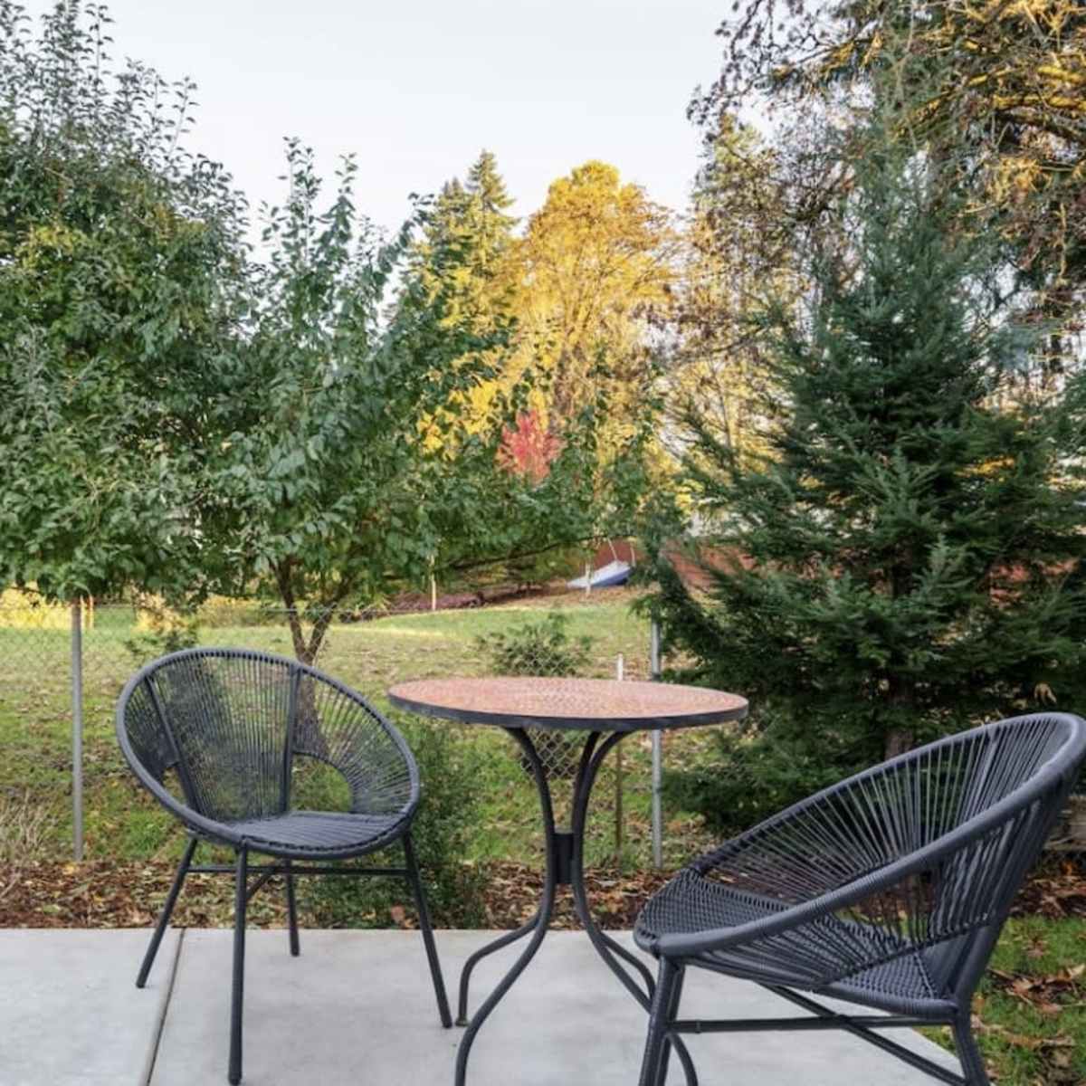 A small patio features two chairs and a round table, with a backdrop of trees and an open grassy area beyond a fence.