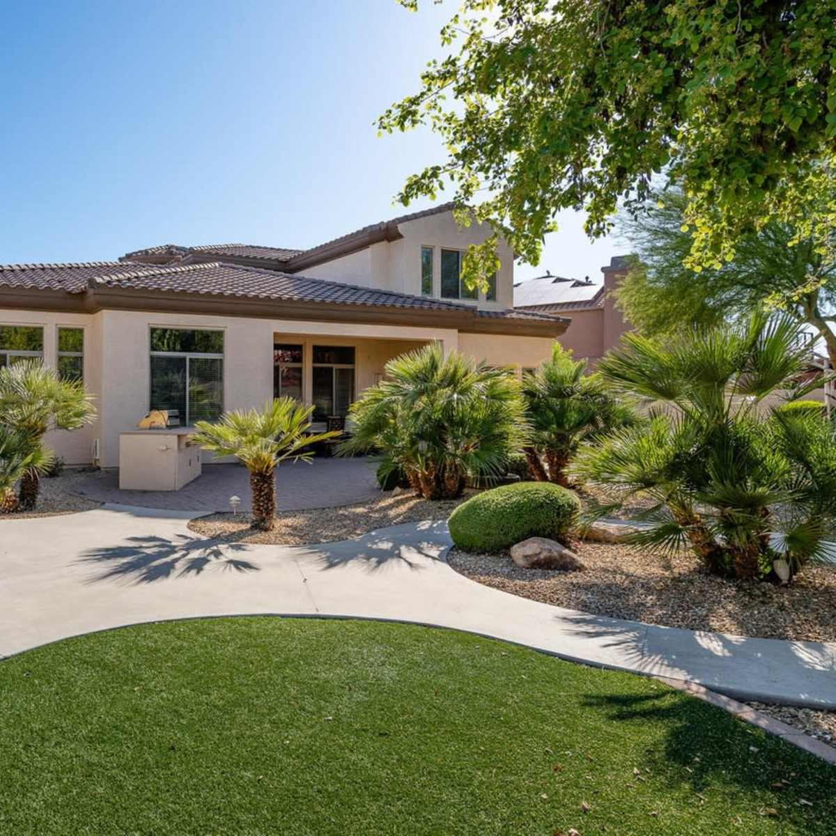 A landscaped backyard features a patio area with seating, surrounded by palm trees and greenery, leading to a two-story house.