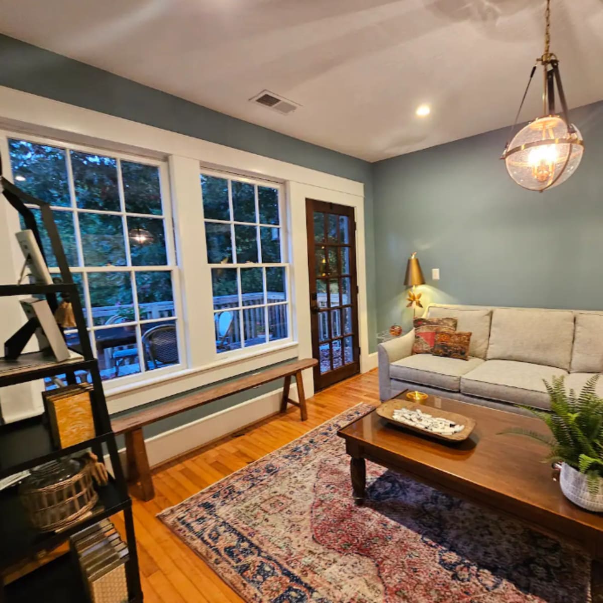 The scene features a cozy living room with a light gray sofa, a wooden coffee table, and large windows allowing natural light to illuminate the space.
