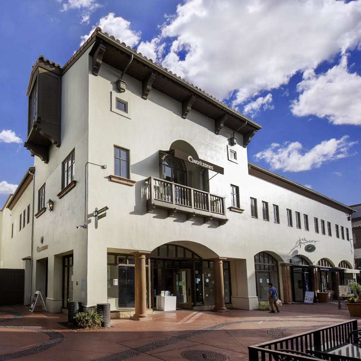 A two-story building with a balcony, located at the corner of a pedestrian plaza under a blue sky with scattered clouds.
