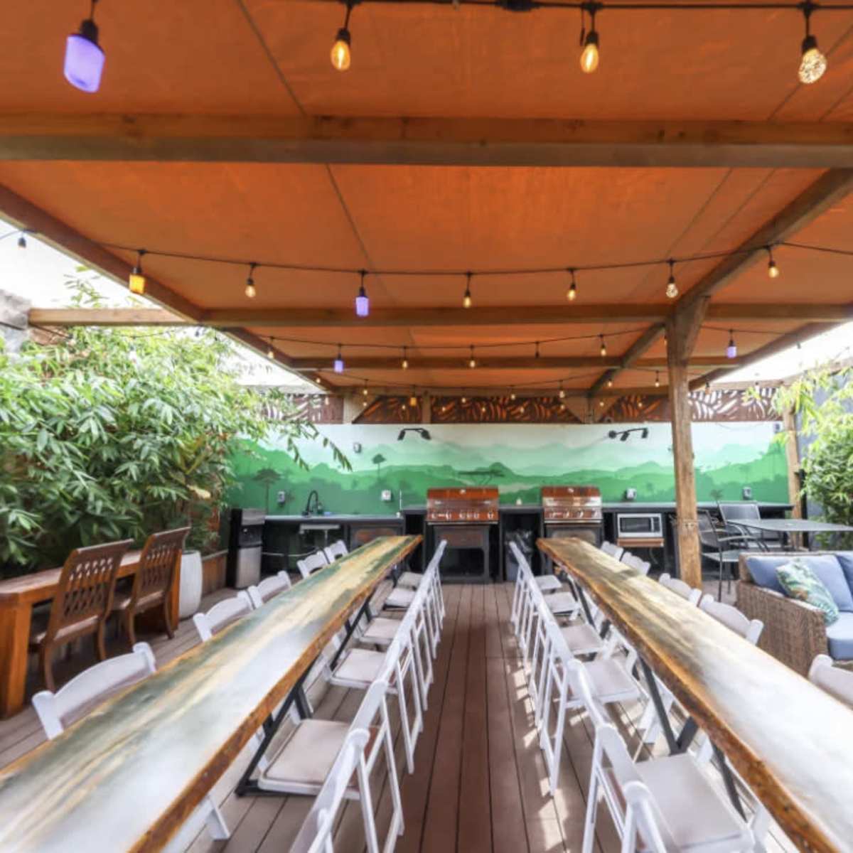 An outdoor dining area with long tables, chairs, and a seating arrangement under a canopy of lights and a painted mural in the background.