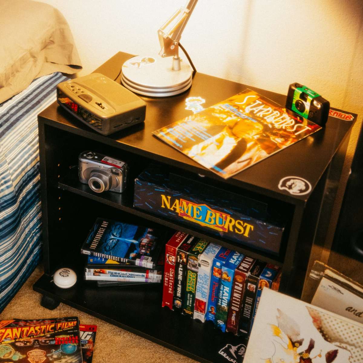 A bedside table holds various items, including magazines, a film camera, and VHS tapes, next to a neatly made bed with a striped comforter.