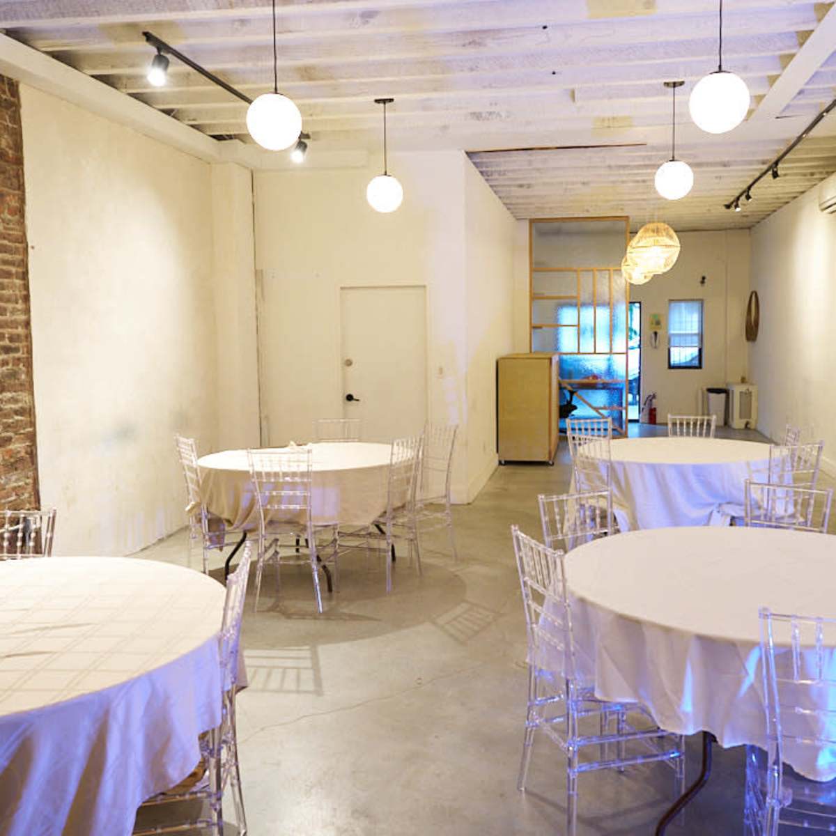 A spacious event room features several round tables covered with white tablecloths and illuminated by pendant lights.