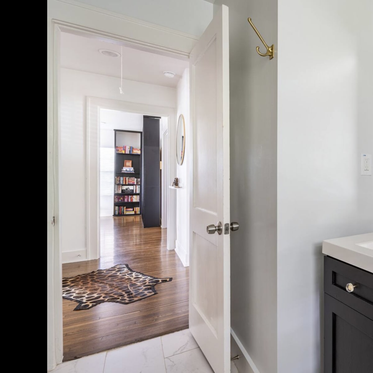 A bathroom vanity with a gold faucet and a large mirror, leading to a hallway with bookshelves visible in the background.
