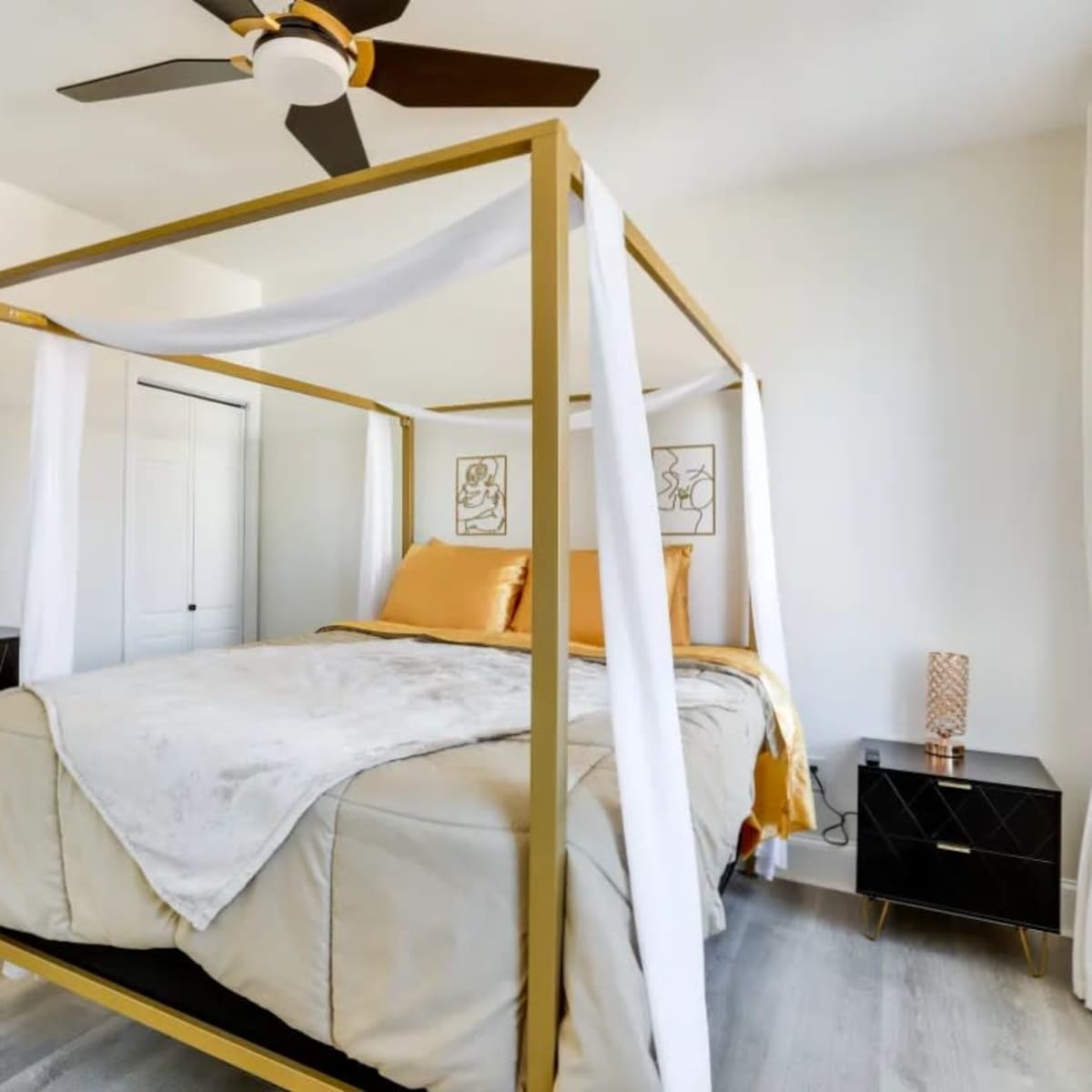 A gold four-poster bed with sheer white draping is positioned in a bright room with light-colored walls and modern black nightstands.