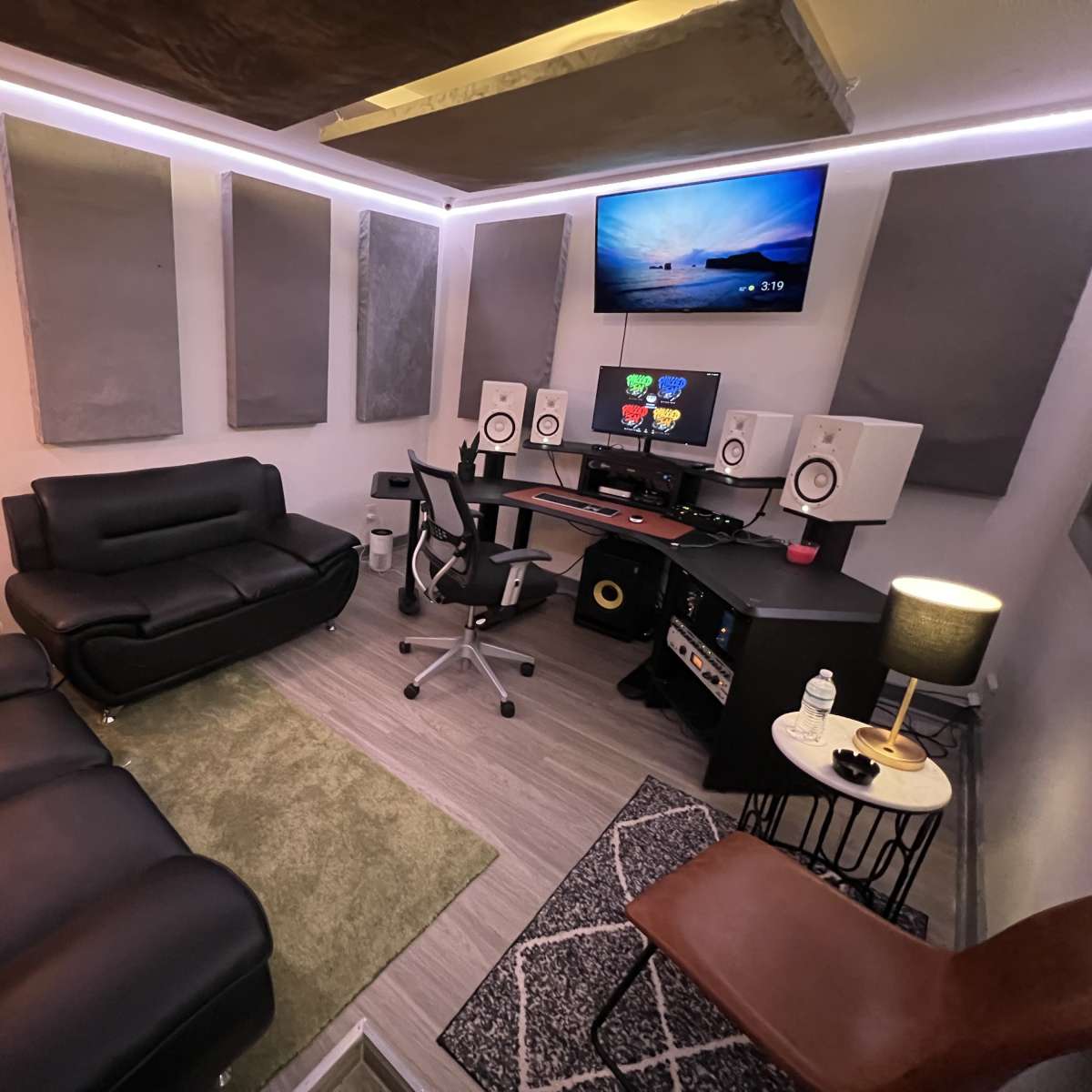 A compact music studio featuring a black sofa, a desk with speakers and a computer monitor, soundproofing panels on the walls, and various seating arrangements.
