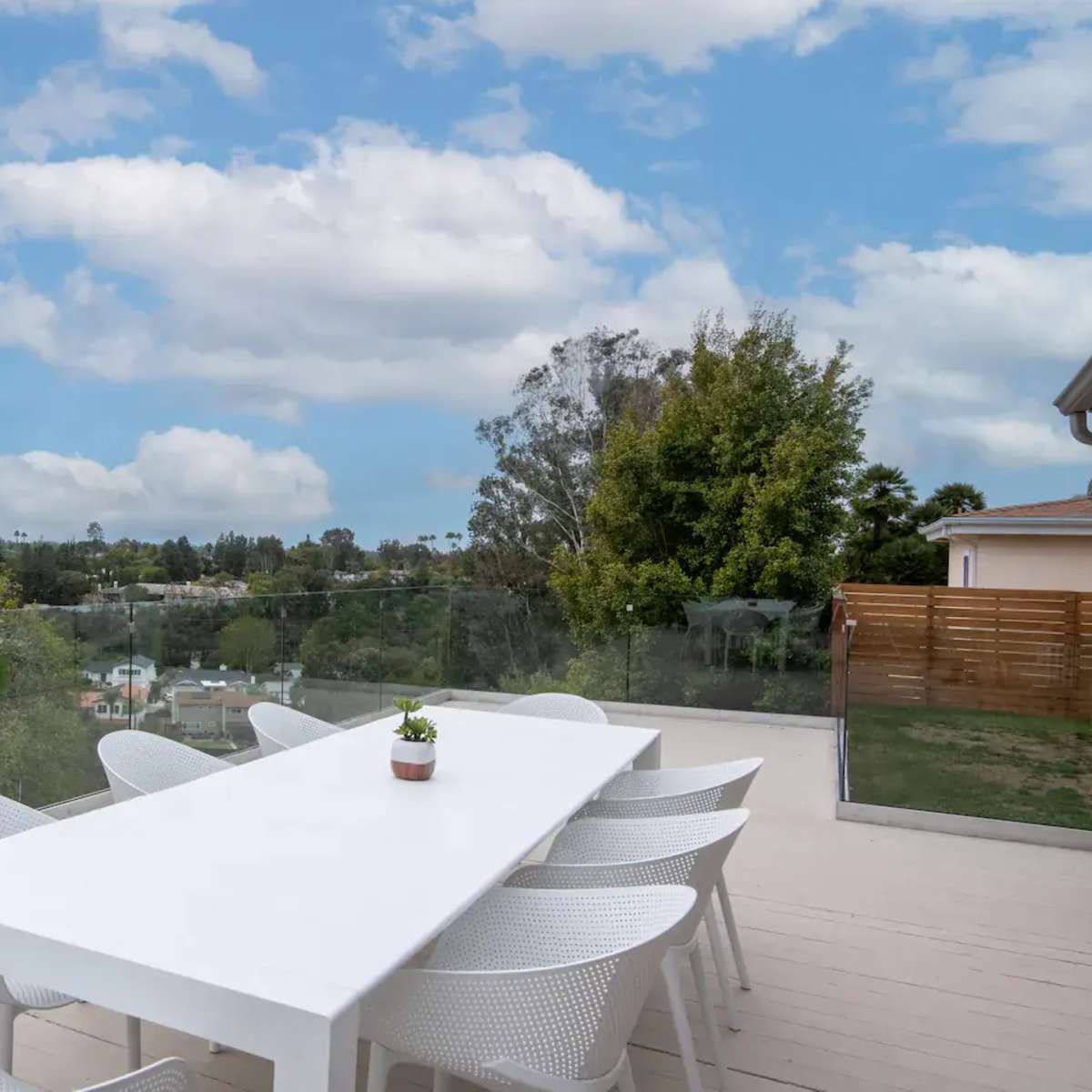 A modern outdoor dining area features a long white table surrounded by chairs, with a view of greenery and a blue sky in the background.
