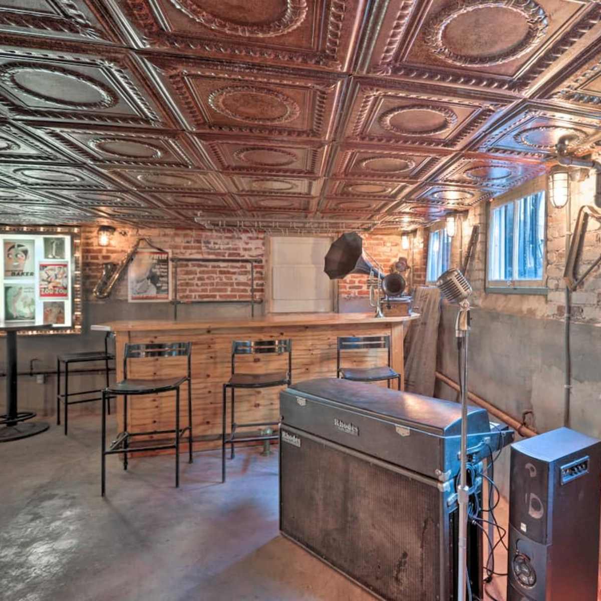 A basement bar area with a tin ceiling, exposed brick walls, and vintage-style microphones set up near a wooden counter and seating.