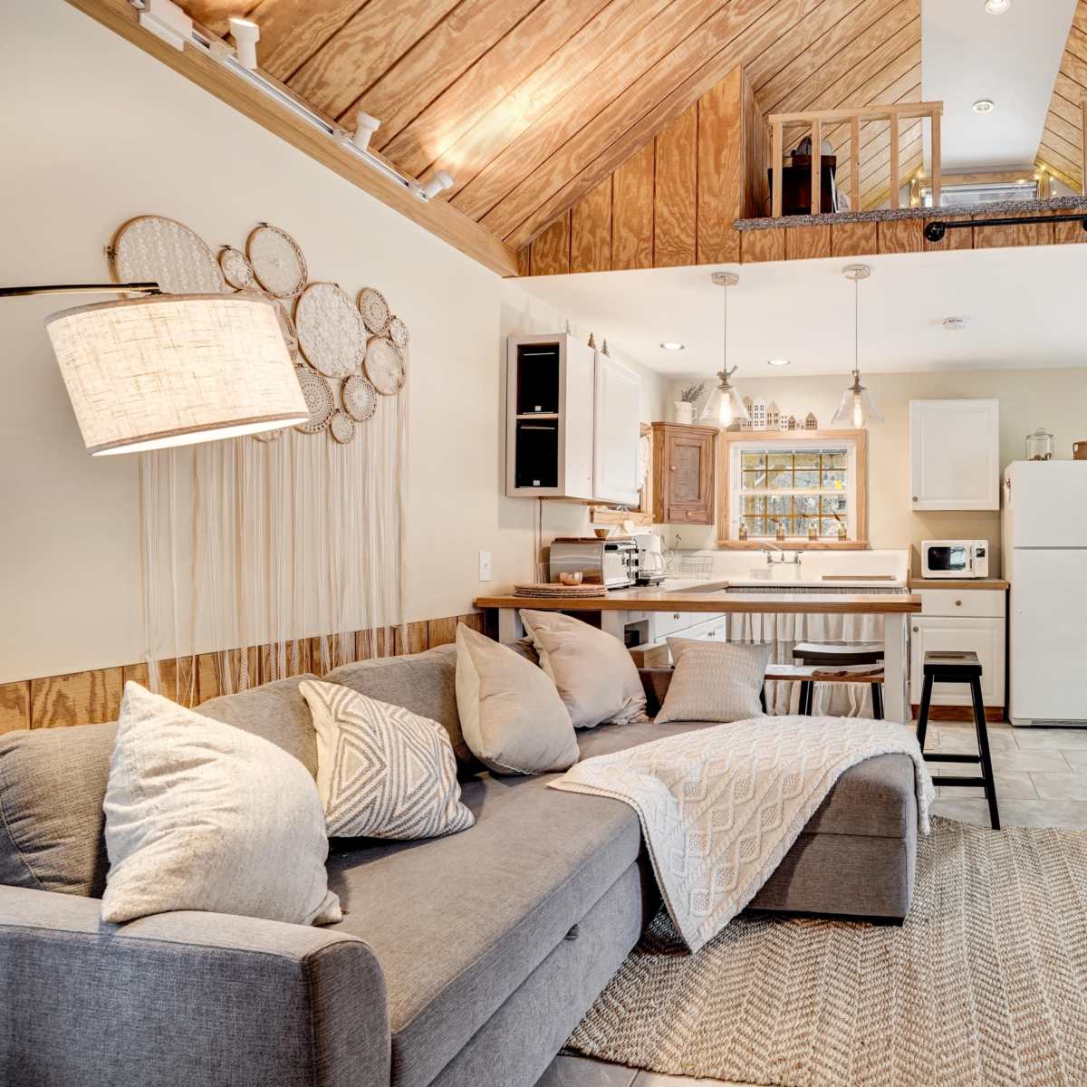 A cozy living space with a gray sofa, a kitchen area including a refrigerator and microwave, and wooden accents highlighted by a lofted ceiling with exposed beams.