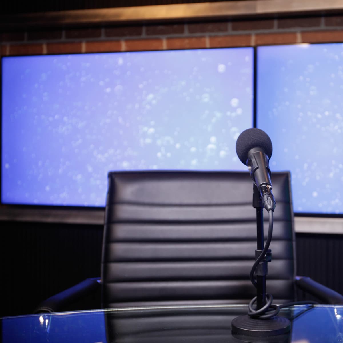 A black microphone stands in front of an empty chair at a glass table, with two large screens displaying a blue background behind them.