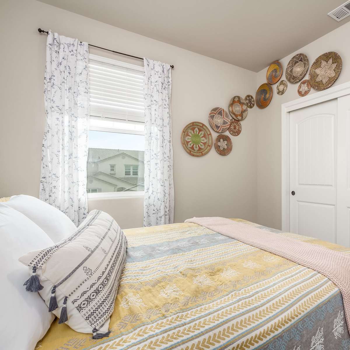 A cozy bedroom featuring a patterned bedspread, decorative pillows, and woven wall art above a window with sheer curtains.