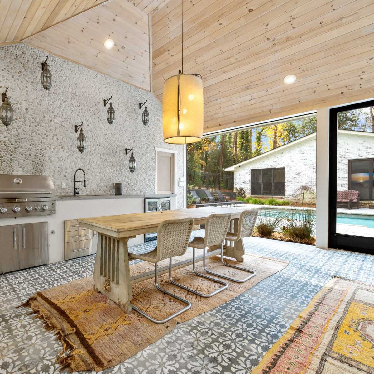 A modern outdoor kitchen area featuring a stainless steel grill, a wooden dining table with metal chairs, and decorative tile flooring.