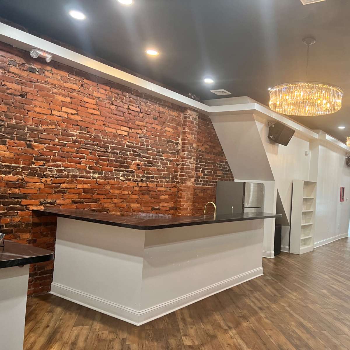 An interior space featuring a brick wall, a modern bar counter, a small refrigerator, and chandeliers hanging from the ceiling, with white drapes along one side.