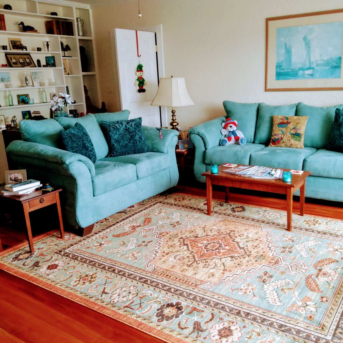 A light-colored living room features two turquoise sofas, a wooden coffee table, and a decorative rug, with built-in shelves in the background.