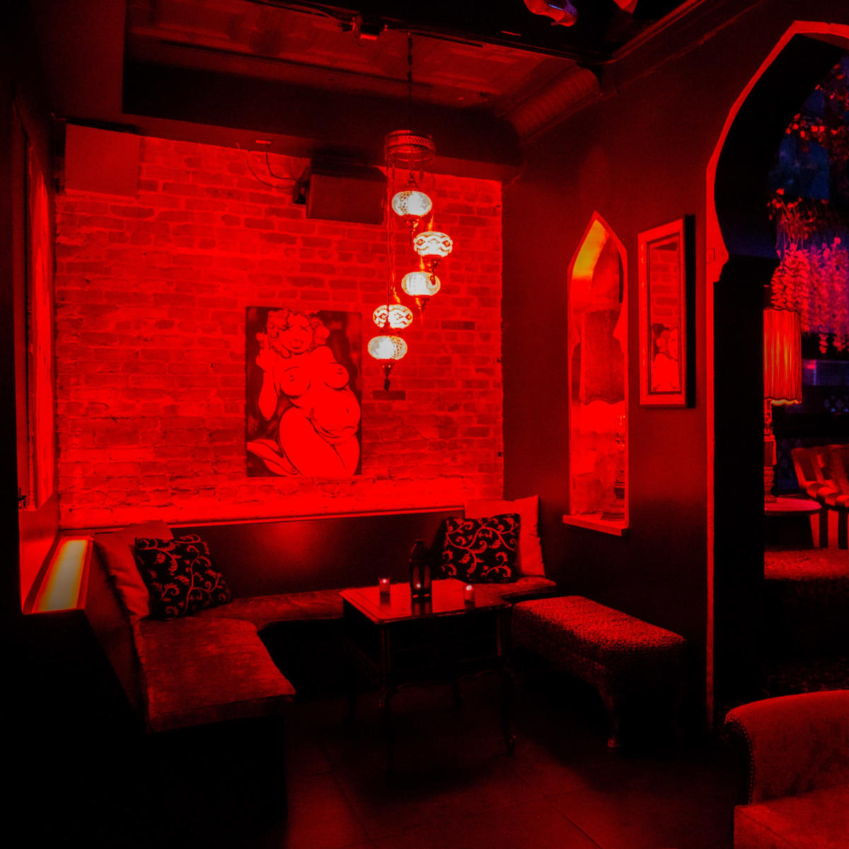 A dimly lit lounge with red lighting, featuring a plush sofa, a small table, and an artwork on the brick wall.
