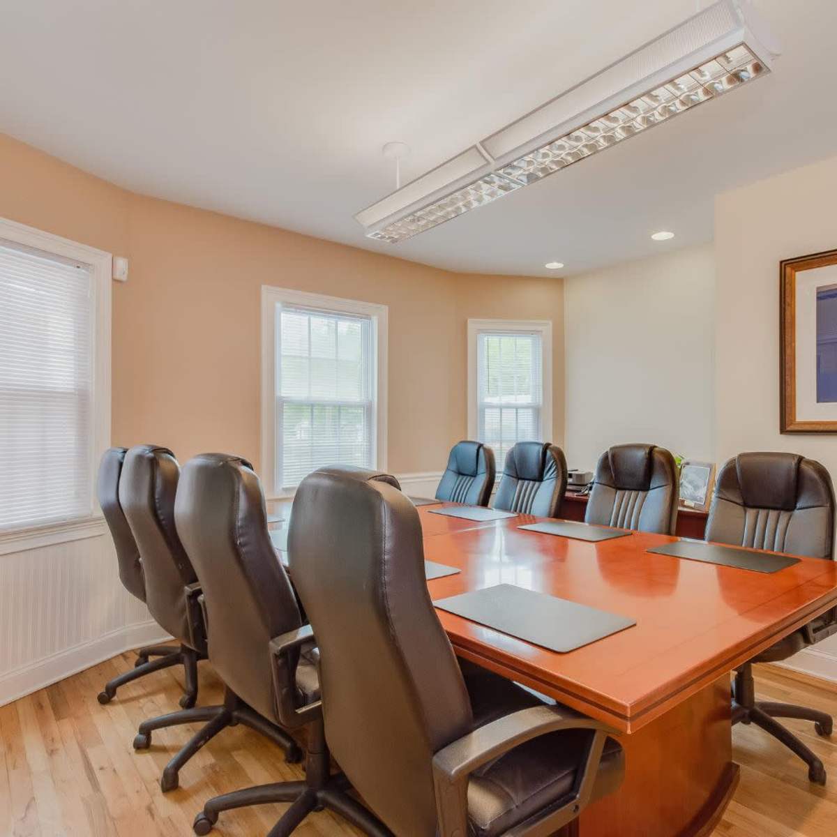 A conference room with a large wooden table surrounded by eight black leather chairs and several windows allowing natural light.