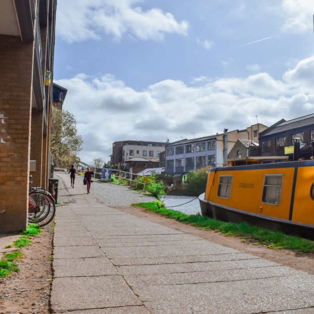 A yellow narrowboat moored alongside a paved walkway next to a canal, with modern buildings in the background and people walking along the path.
