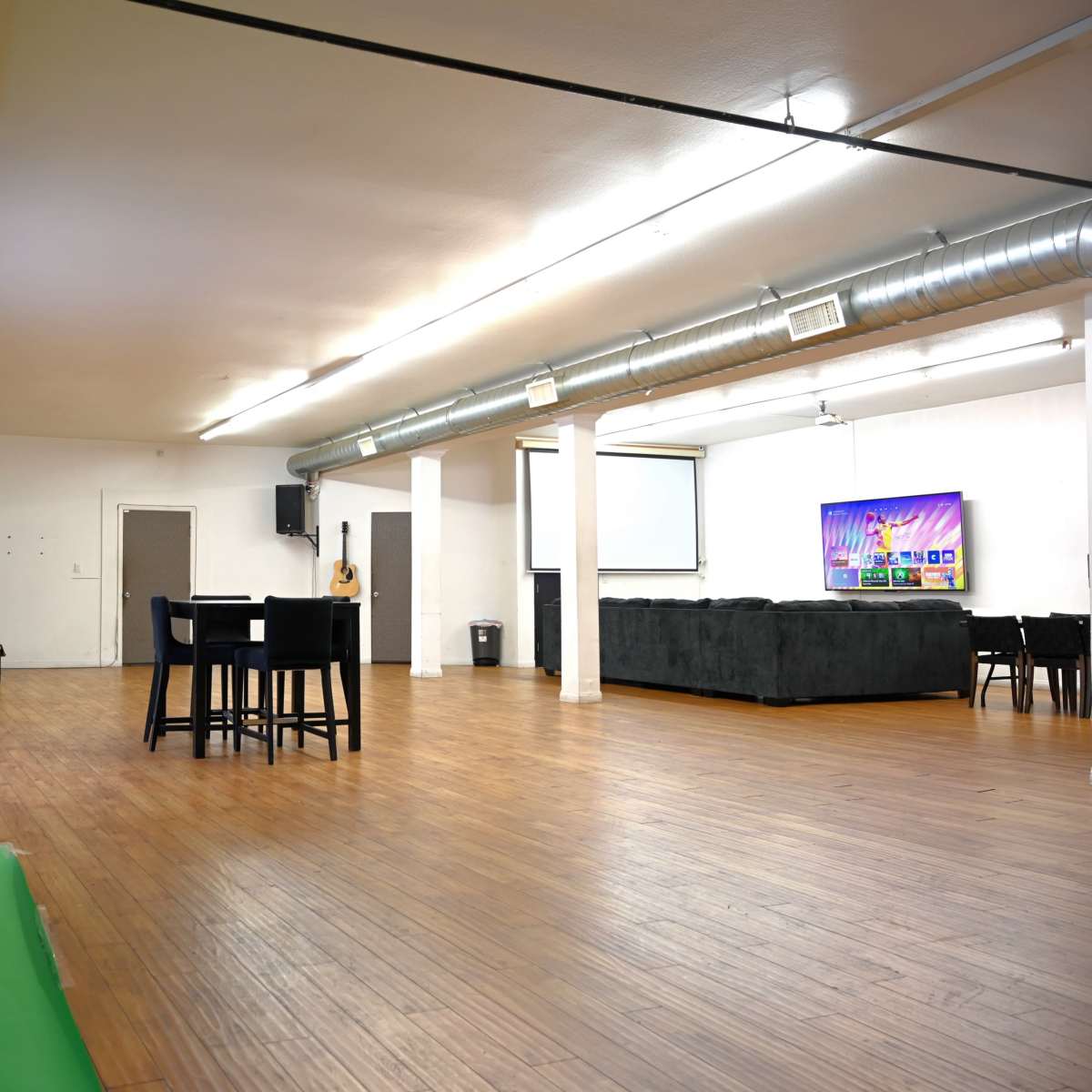 A spacious, empty room featuring a green backdrop on the left, a seating area, and a table with chairs in the center, along with a screen and a couch in the background.