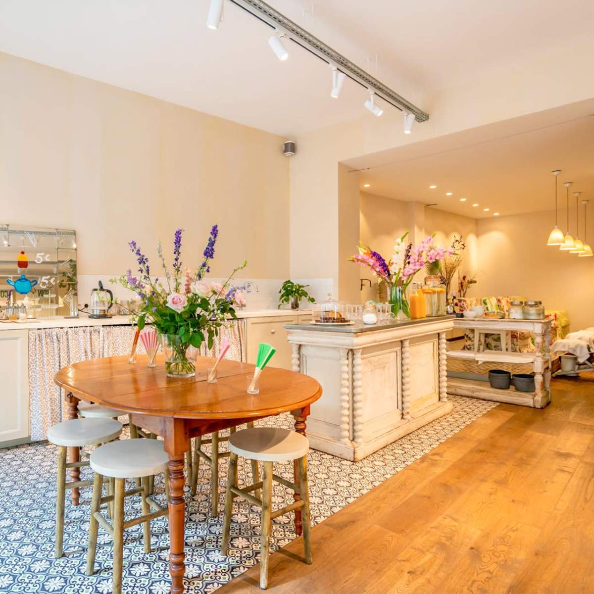 A bright kitchen and dining area features a round wooden table surrounded by chairs, with floral arrangements and a side counter displaying food and drinks.